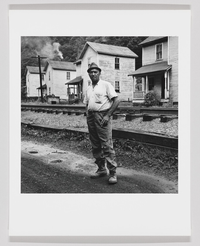 A man wearing a hat stands by railroad tracks in front of wooden houses.
