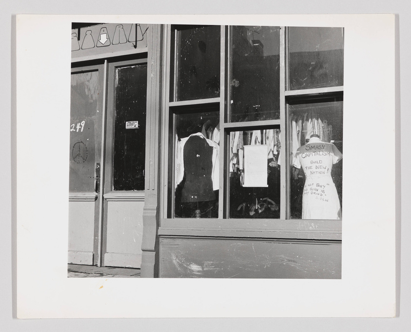 Black and white photo of a storefront with a display window showing a T-shirt with the slogan "SMASH CAPITALISM BUILD THE NEW NATION" and other clothing items. Signs with protest messages are visible, and the store's entrance has the number 249 above the door.