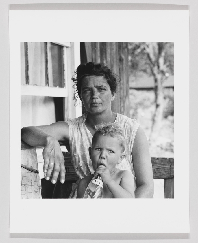 A black and white photograph of a woman and a young child sitting together. The woman, with her hair tousled and a serious expression, is leaning on a wooden railing with one arm draped over it. The child, who is in front of her and appears to be sucking on the top of a glass bottle, is looking directly at the camera with a slightly furrowed brow. They are both in a shaded area with a window and part of a house visible in the background.