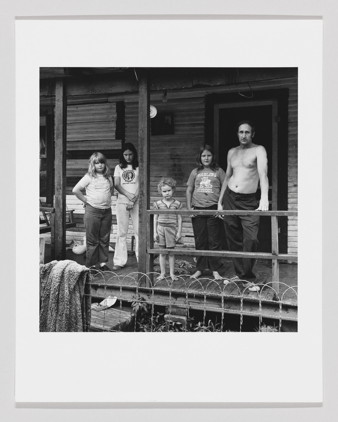 A black and white photograph of a family of five standing on a wooden porch. From left to right, there are three children and two adults. The children are wearing casual clothes, with two girls flanking a younger boy in the center. The man on the right is shirtless and wearing pants, leaning slightly on the porch railing. The wooden structure of the house appears weathered, and there's a towel hanging on the railing to the left. The family gazes directly at the camera with neutral expressions.