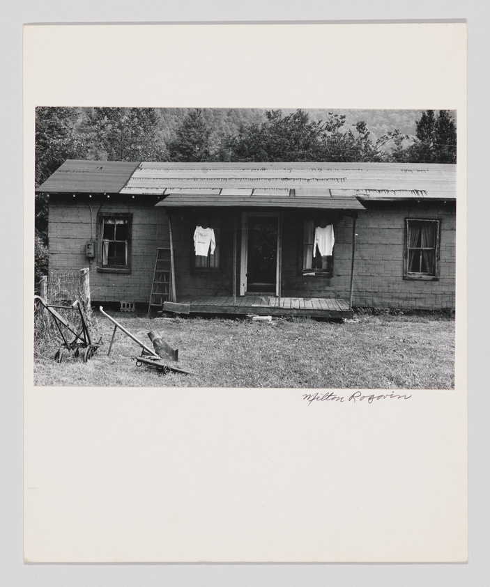 A black and white photograph of a modest, single-story wooden house with a front porch. Clothes are hanging on a line to dry, and there's an old-fashioned push mower and a ladder on the unkempt lawn. Trees can be seen in the background, suggesting a rural setting. The image is signed "Milton Rogovin" at the bottom right.