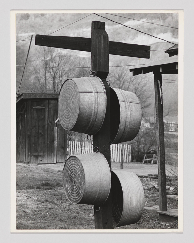 A black and white photograph showing three metal washbasins hanging vertically on a wooden post, with a rural backdrop including a wooden shed and bare trees.