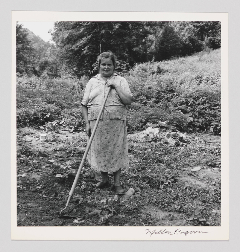 A black and white photograph of a woman standing in a garden with a hoe in her hand. She is wearing a patterned skirt and a short-sleeved blouse, and she looks directly at the camera with a slight smile. The garden appears to be in a rural setting with dense foliage in the background. There is a signature at the bottom right of the image.