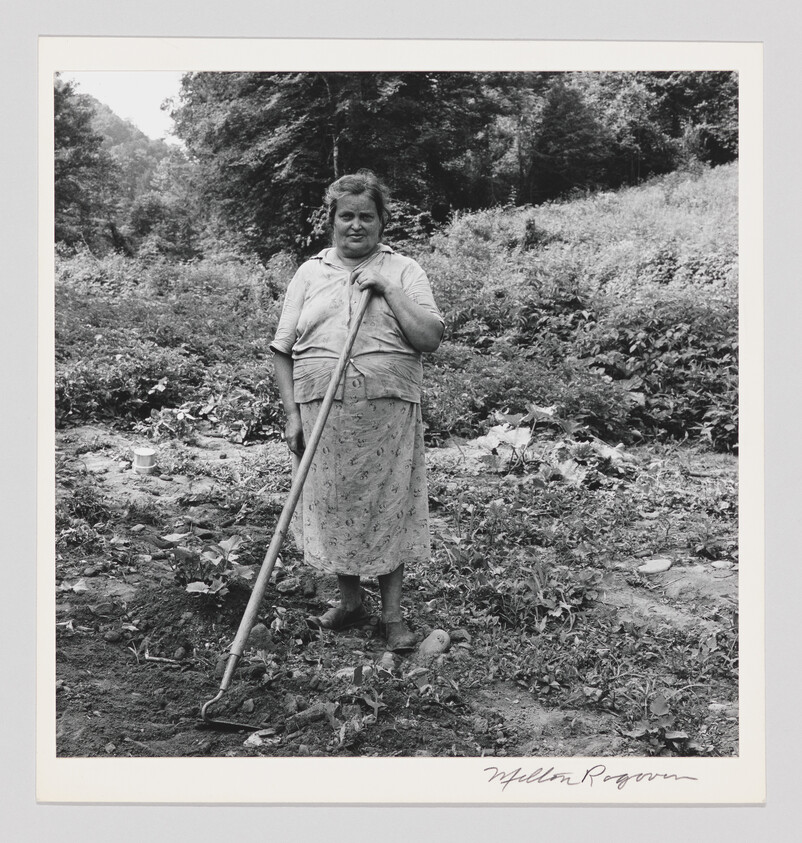 An older woman stands in a garden holding a hoe, surrounded by plants and dirt.
