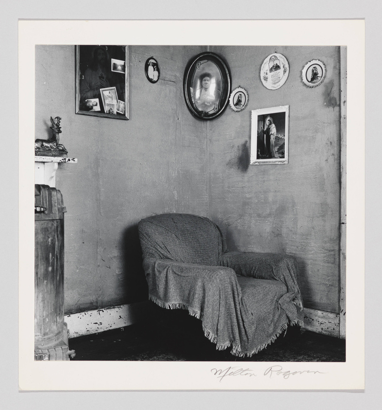 A black and white photograph depicting a corner of a room with a worn wall adorned with various framed pictures, including portraits and decorative plates. A single armchair covered with a fringed throw blanket sits below the images, and a small figurine rests on a shelf to the left. The photo is signed by the photographer at the bottom right.