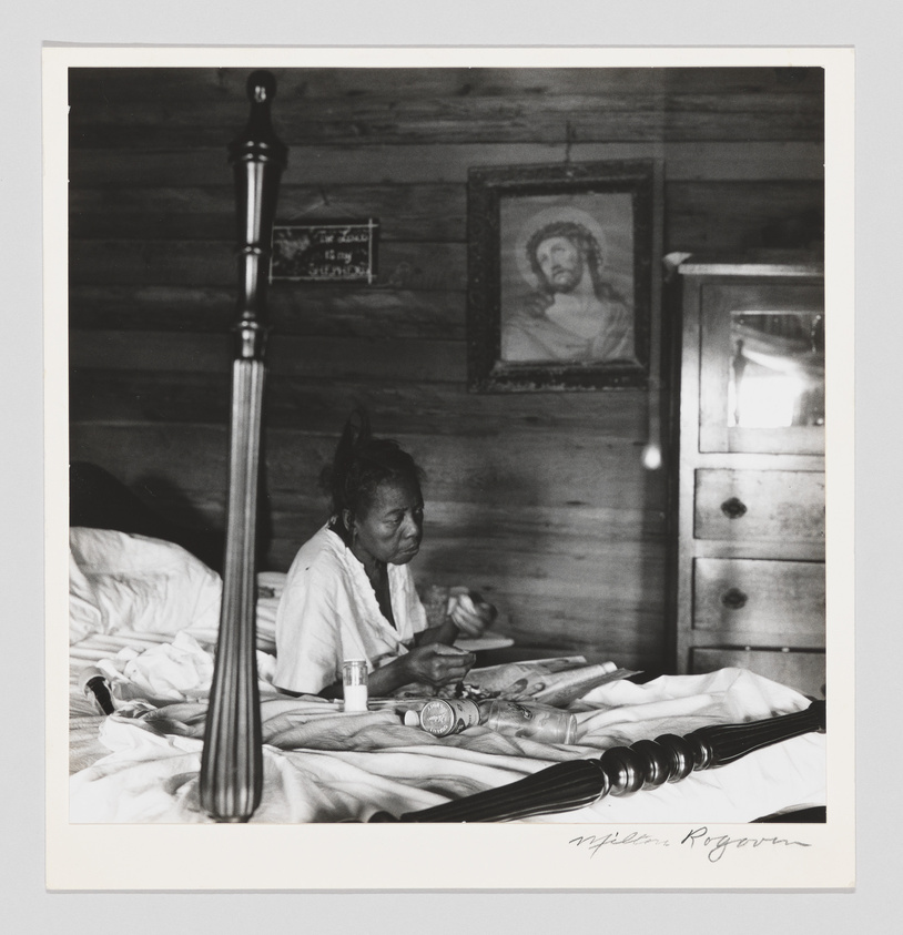 A black and white photograph depicting an elderly woman sitting up in bed, holding what appears to be a small object in her hands. The room has wooden walls, and there is a framed picture of Jesus on the wall above a wooden cabinet. The bed has a turned wooden bedpost in the foreground, and there are various items, including a bottle, on the bedspread. The image includes a signature at the bottom right corner.