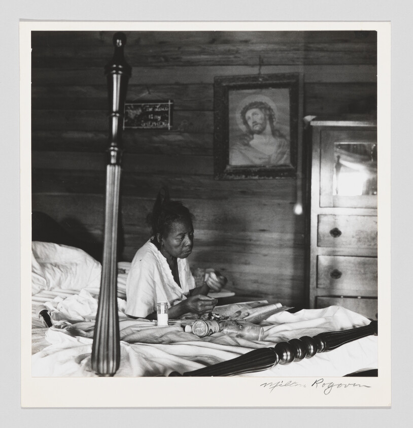 An older woman sits on a bed sorting medicine bottles with a framed Jesus portrait on the wall.