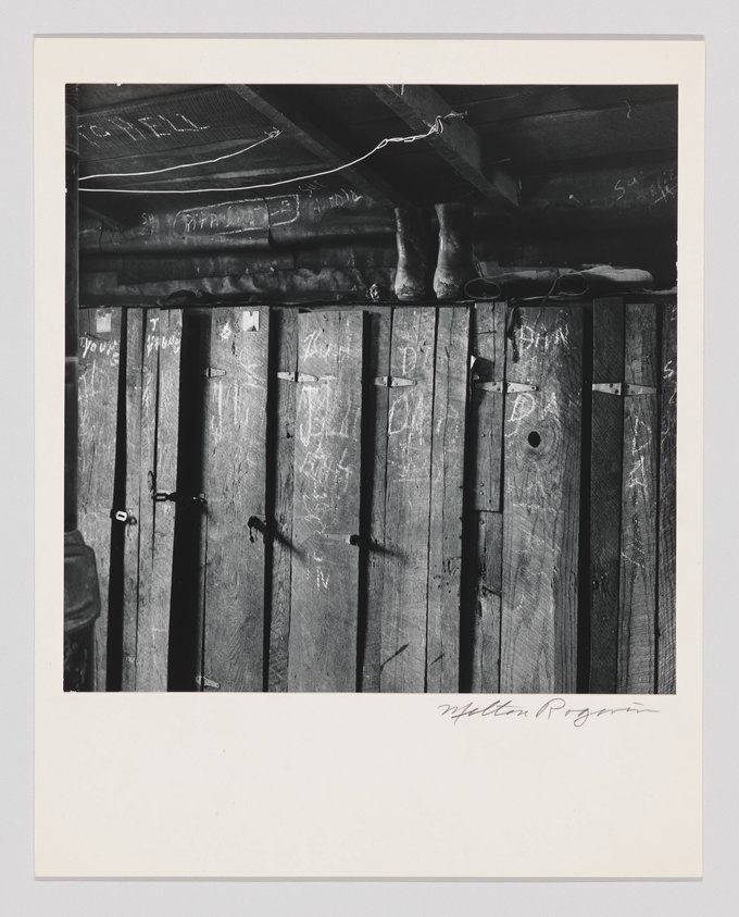 Black and white photograph depicting a rustic wooden interior with vertical planks, some of which are inscribed with graffiti. A pair of boots is perched on a horizontal beam above the planks, and a wire hangs loosely from the ceiling. The image is framed with a white border, and there is a signature at the bottom right corner.