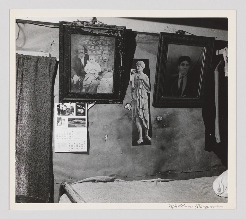 A black and white photograph depicting a wall with framed images, including a family portrait, a classical statue, and a portrait of a man. A calendar hangs on the left side, and there's a bed in the foreground with a pillow and blanket. The wall appears aged with some peeling and damage. The photo is signed at the bottom right.