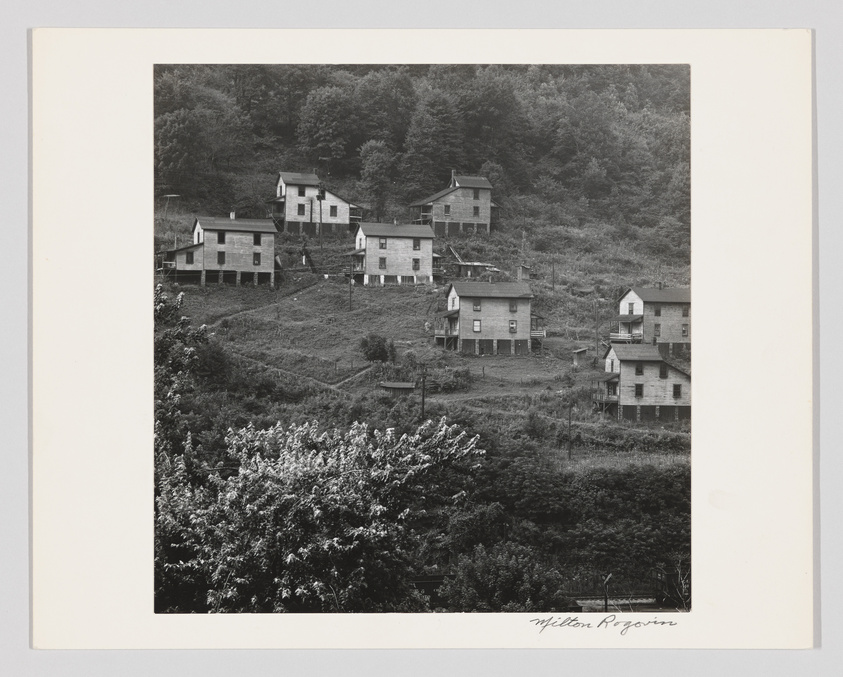 A black and white photograph depicting a hillside scattered with a collection of old, weathered houses, some with visible wear and tear. The houses are surrounded by trees and overgrown vegetation, suggesting a rural or semi-abandoned setting. The image has a signature in the lower right corner that reads "Milton Rogovin."