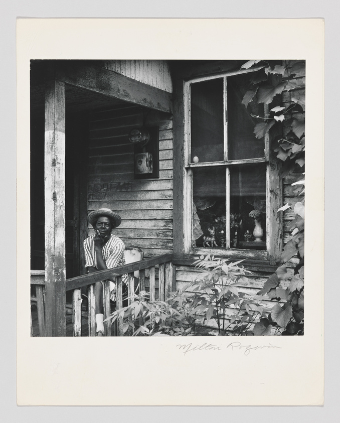 A black and white photograph of an individual wearing a striped shirt and a hat, standing on the porch of a weathered wooden house. The person is leaning on the railing, surrounded by lush foliage and a window with a broken pane next to them. The image has a signature at the bottom right corner.