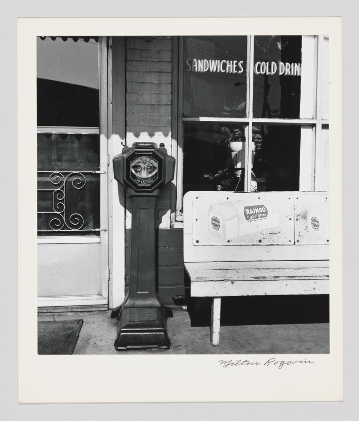 A black and white photograph depicting a vintage parking meter in the foreground with a clear view of its coin slot and timer. Behind the meter, part of a storefront is visible, featuring a window with the words "SANDWICHES COLD DRIN" painted on it, suggesting "COLD DRINKS" with the last letter cut off. The window displays a reflection and some items inside the store. To the right, there's a bench with a "RAMBO" sticker on it. The image has a white border with the artist's signature, "Milton Rogovin," at the bottom right.