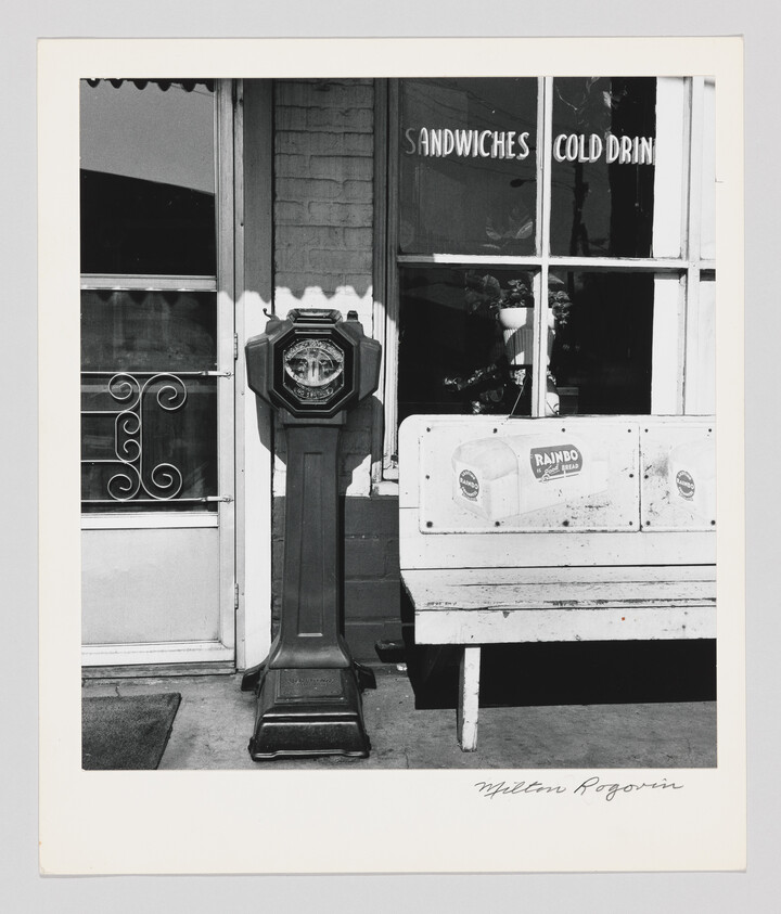 An old coin-operated standing scale sits outside a sandwich shop beside a white bench.