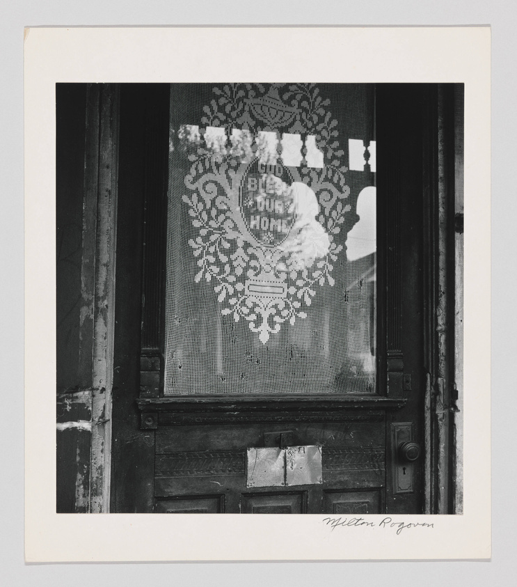 A black and white photograph of a weathered door with a lace curtain featuring the phrase "God Bless Our Home" visible through the glass pane. The door has a mail slot and is framed within a dark entryway, with the artist's signature, Milton Rogovin, at the bottom right on the matting.