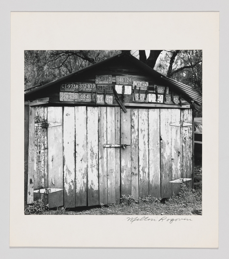 Black and white photograph of a weathered wooden shed with a collection of old license plates affixed to its side, surrounded by a natural environment with trees in the background. The image is signed "Milton Rogovin" at the bottom.