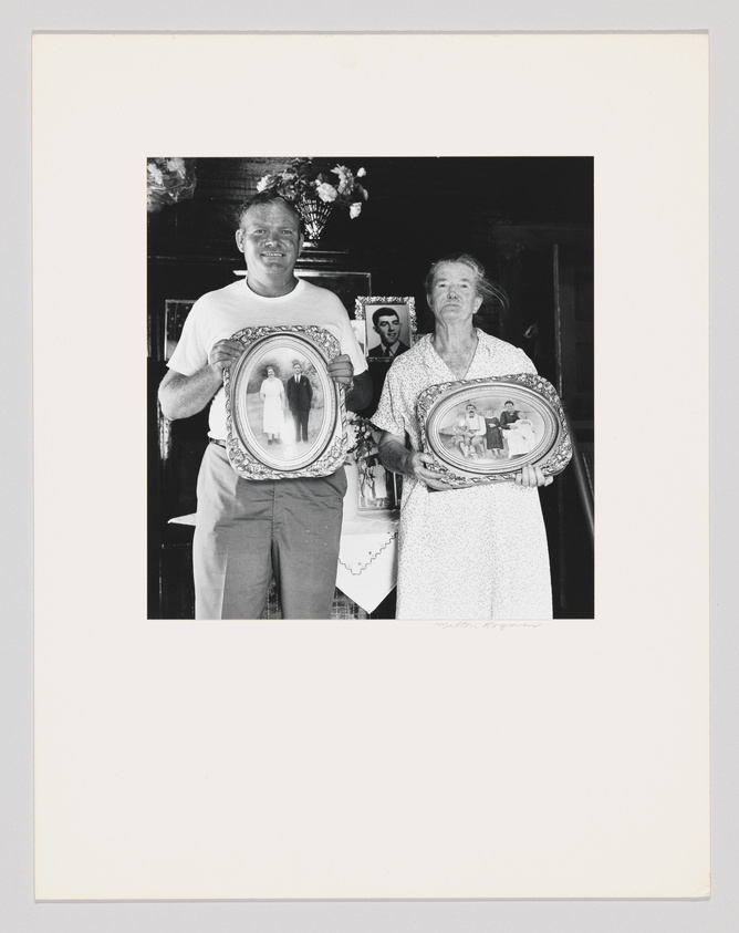 A black and white photograph of a smiling man and a woman standing side by side, each holding an ornate oval-framed portrait. The man on the left holds a portrait of a couple, while the woman on the right holds a portrait of two children. Behind them is a wall with a floral arrangement and another framed picture. The image has a white border with the photographer's signature at the bottom right.