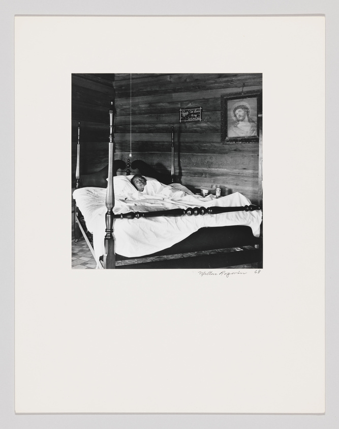 A black and white photograph depicting an elderly person lying in a traditional four-poster bed in a rustic room with wooden walls. A framed portrait of a figure hangs on the wall above a small, framed religious text. The bedding is white and contrasts with the dark wood of the bed and walls. The image includes a signature at the bottom right corner.