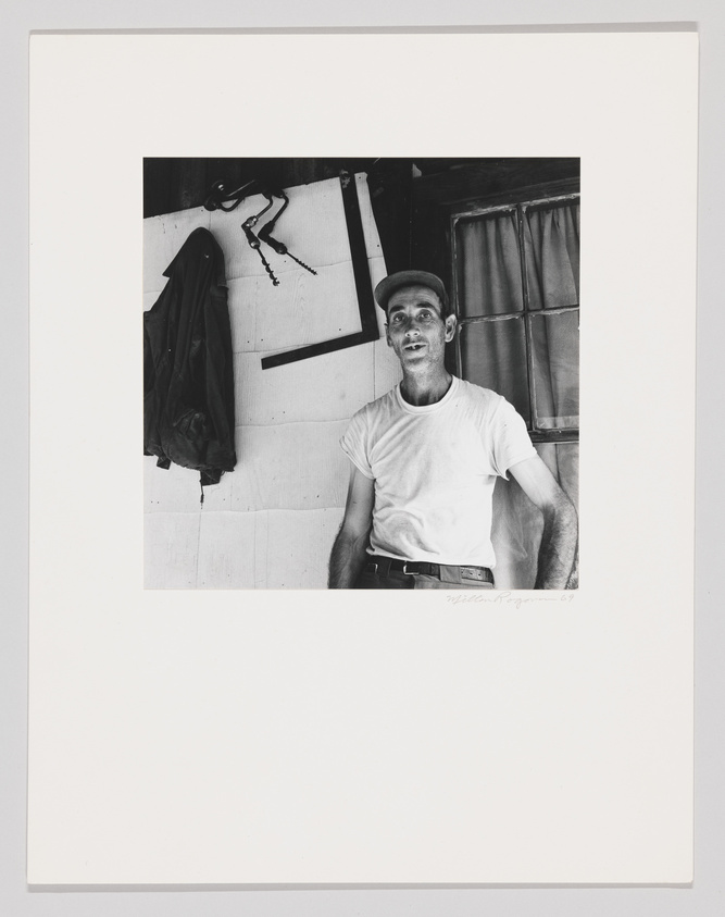 Black and white photograph of a man wearing a cap and a white T-shirt, standing in a room with a window and a jacket hanging on the wall. The man is looking upwards with a slight smile, and there is a signature at the bottom right of the image.