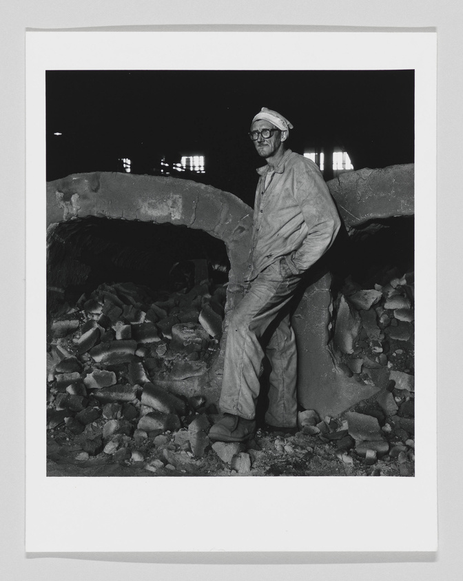 A black and white photograph of a worker wearing a hard hat, glasses, and overalls, standing amidst rubble and leaning against a large broken concrete pipe. The background is dark with a few illuminated windows in the distance.