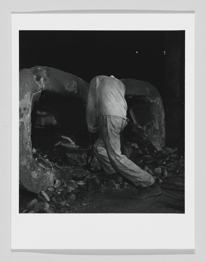 A worker leans into a large industrial furnace, removing debris from the opening.