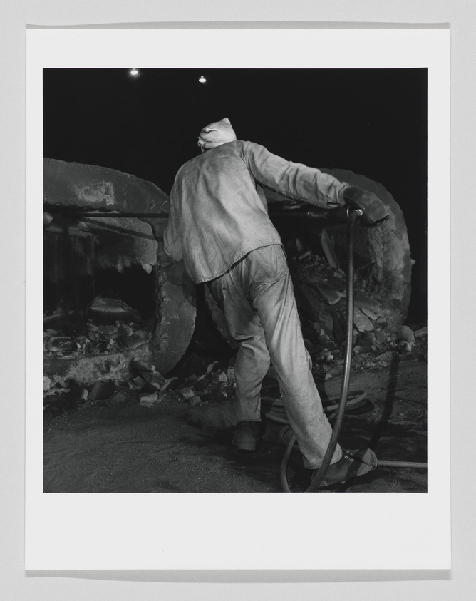 A black and white photograph of a worker in a hard hat and coveralls pushing a large wheelbarrow in an industrial setting, possibly a construction site or mine. The lighting is dim, and the focus is on the person's action and the rough texture of the materials around them.