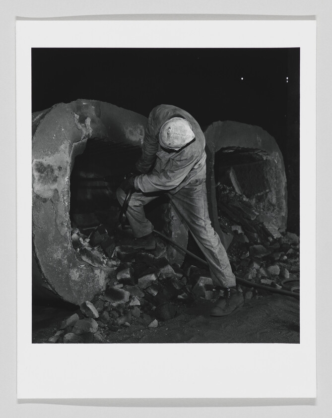 A worker wearing protective clothing breaks apart large concrete pipes with a pry bar at night.