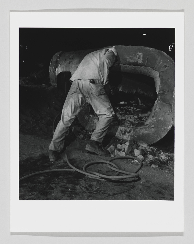 A black and white photograph capturing a worker bent over while working on a large, rugged tire of heavy machinery at night. The worker's face is not visible, and the environment suggests an industrial or construction setting. A hose lies on the ground near the worker's feet.