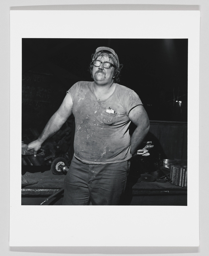 A black and white photograph of a man standing confidently with his hands on his hips in a workshop environment. He is wearing a dirty T-shirt, work trousers, and a cap with protective goggles pushed up on his forehead. His shirt is stained with what appears to be dust or soot, suggesting he may be involved in some kind of manual labor or craftsmanship. The background is indistinct, but there are hints of tools and machinery, reinforcing the industrial setting.