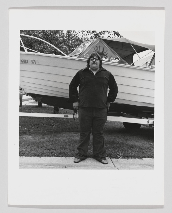 A black and white photograph of a person standing in front of a boat on a trailer. The individual is wearing glasses, a dark long-sleeve shirt, and pants, with their hands at their sides. Trees and a grassy area can be seen in the background.