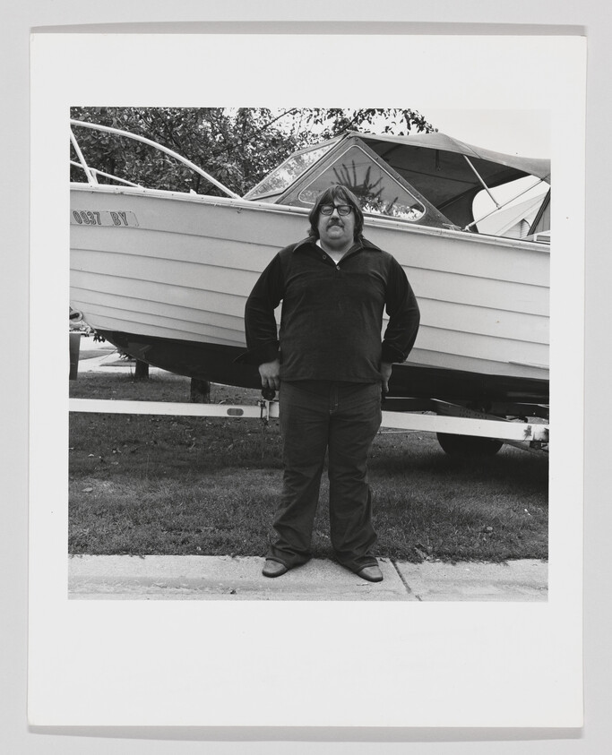 A man stands in front of a boat on a trailer parked on a lawn.