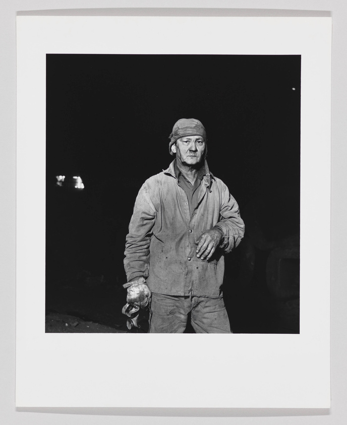 A black and white photograph of a man standing in the foreground, looking directly at the camera. He appears to be a worker, wearing a dirty long-sleeve shirt, pants, and a cap, with a headlamp attached to it. He is holding gloves in his left hand and has a serious expression on his face. The background is dark, with only a faint hint of light, suggesting an industrial or underground setting.