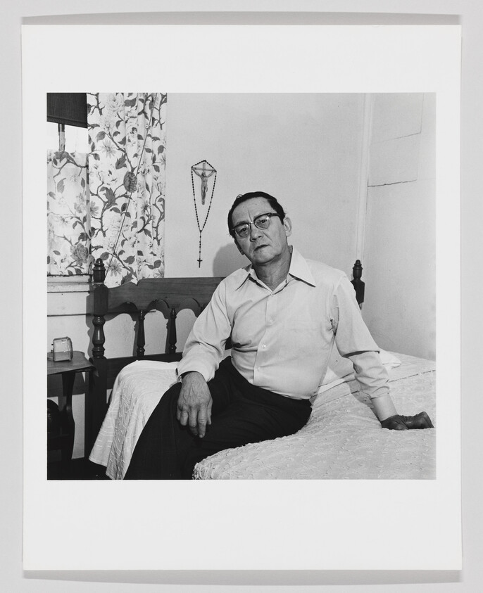 A middle-aged man wearing glasses sits on a bed in a simple room with a rosary on the wall.