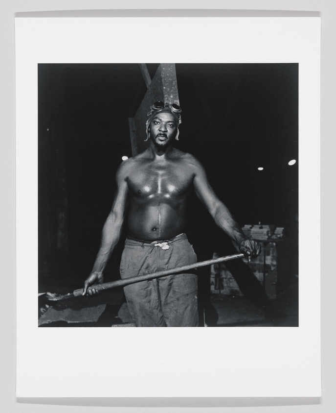 A black and white photograph of a shirtless man wearing goggles on his head and holding a long pole across his body. He appears to be in a workshop or industrial setting.