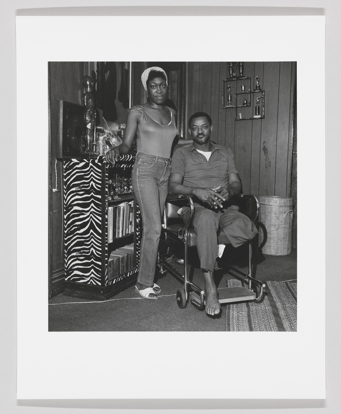 A black and white photograph of a woman standing and a man seated in a vintage interior setting. The woman is wearing a sleeveless top and jeans, with her hand resting on a bar cart adorned with a zebra print cloth. The man is seated in a rolling office chair, wearing a short-sleeved shirt and trousers, with one hand on his thigh and the other on the armrest. Behind them is a wooden cabinet with glassware and a wall-mounted shelf with decorative items. The room has a retro aesthetic with a patterned rug on the floor.