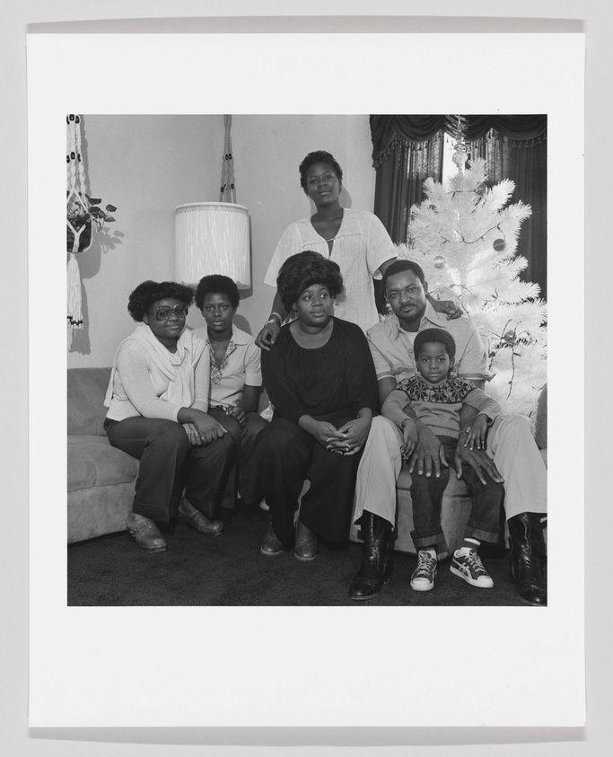 A black and white photo of seven people posing for a family portrait in a living room with a white Christmas tree in the background. Four women are seated on a couch, one man is seated on an armrest, a young boy is seated on the man's knee, and one woman stands behind the couch. They are all dressed in casual attire and appear to be in a festive mood.