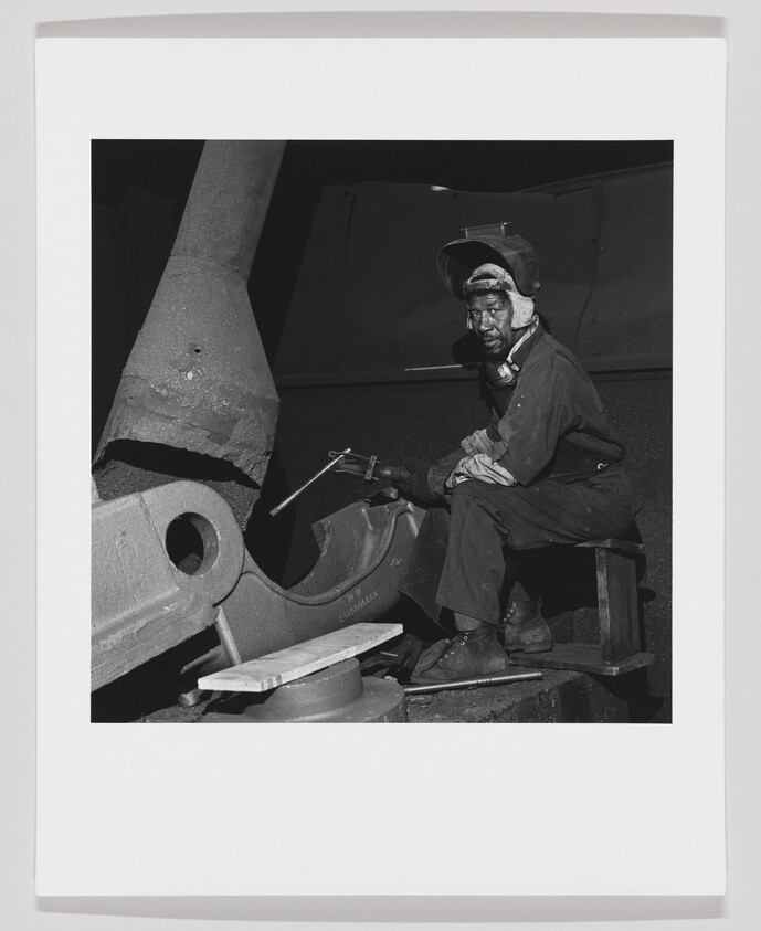 A black and white photograph of a worker sitting on a metal structure, wearing a cap and holding a tool, with large industrial equipment parts surrounding him.