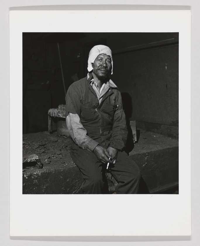 A black and white photograph of a seated man wearing a bandaged head wrap and coveralls, holding a cigarette, with a contemplative expression on his face. The setting appears to be an industrial or workshop environment.