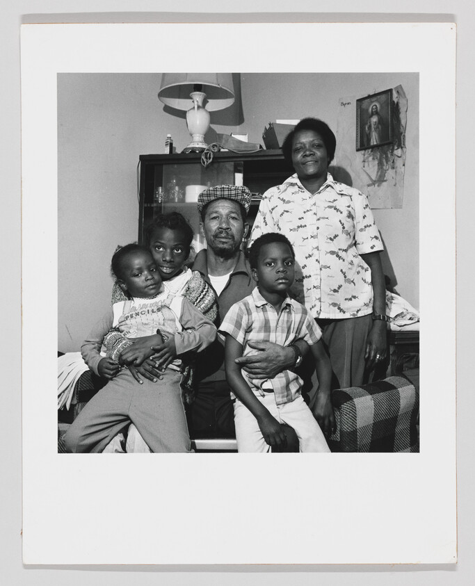 A black and white photograph of a family of five posing together in a living room. A man seated in the center holds a young child on his lap, with another child standing to his right. A woman stands behind the seated man, and another child is held by the man on the left. The room has a simple decor with a religious picture on the wall and a ceiling fan above.
