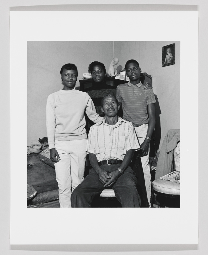 A black and white photograph of four individuals, presumably a family, posing in a modestly furnished room. An elderly man is seated in the center, with two young men standing behind him to the right, and a young woman to the left. A religious icon hangs on the wall, and a stuffed animal sits atop a small cabinet in the background.