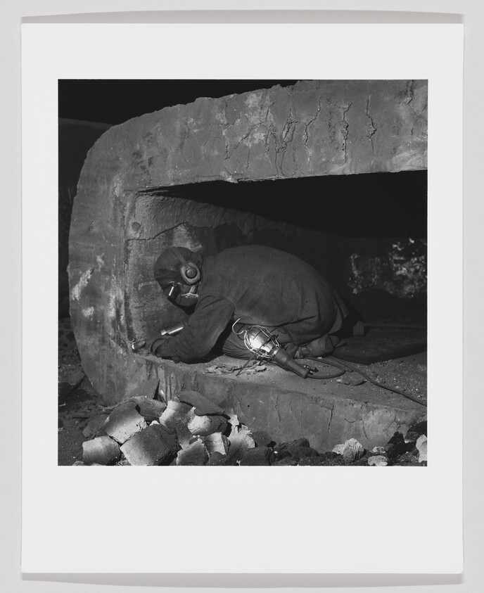 Worker wearing protective mask and headphones crawls into large industrial oven to inspect inside.