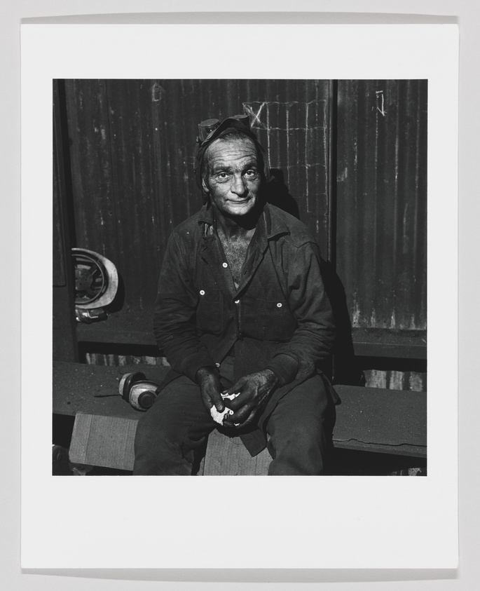 A black and white photograph of an elderly man sitting on a bench with a slight smile on his face. He has noticeable lines and smudges on his face, suggesting he might be a worker or someone who has been involved in physical labor. The man is wearing a dark button-up jacket with the sleeves rolled up, dirty work pants, and has a pair of sunglasses resting on his head. His hands are also dirty, and he is holding a small object in his left hand. The background features a corrugated metal structure, adding to the industrial or work-related theme of the photo.