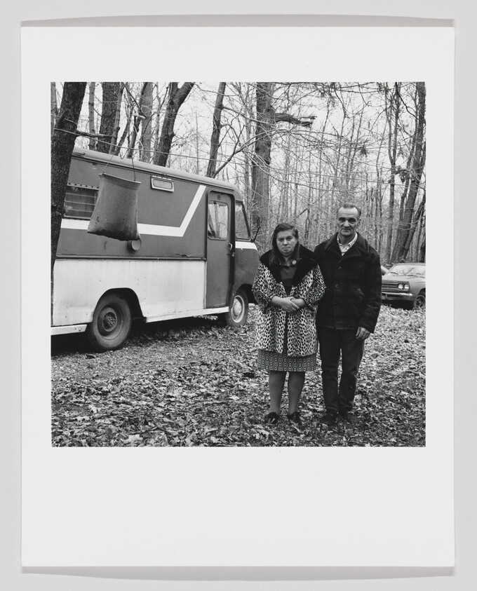 A middle-aged couple stands close together in front of a parked van in a leaf-covered woods.