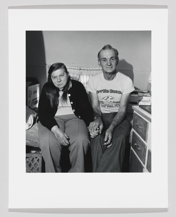 A black and white photograph of an older couple sitting closely together on a bench. The woman on the left is wearing a dark cardigan and light-colored pants, while the man on the right is in a graphic t-shirt and dark pants. Both are looking directly at the camera with neutral expressions, and the setting appears to be a simple, homey interior with a curtain and a small cabinet in the background.