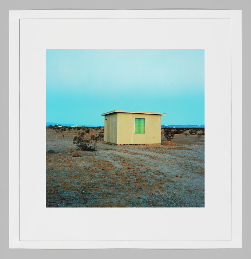 A small yellow shed with a green window stands alone in a flat desert landscape.