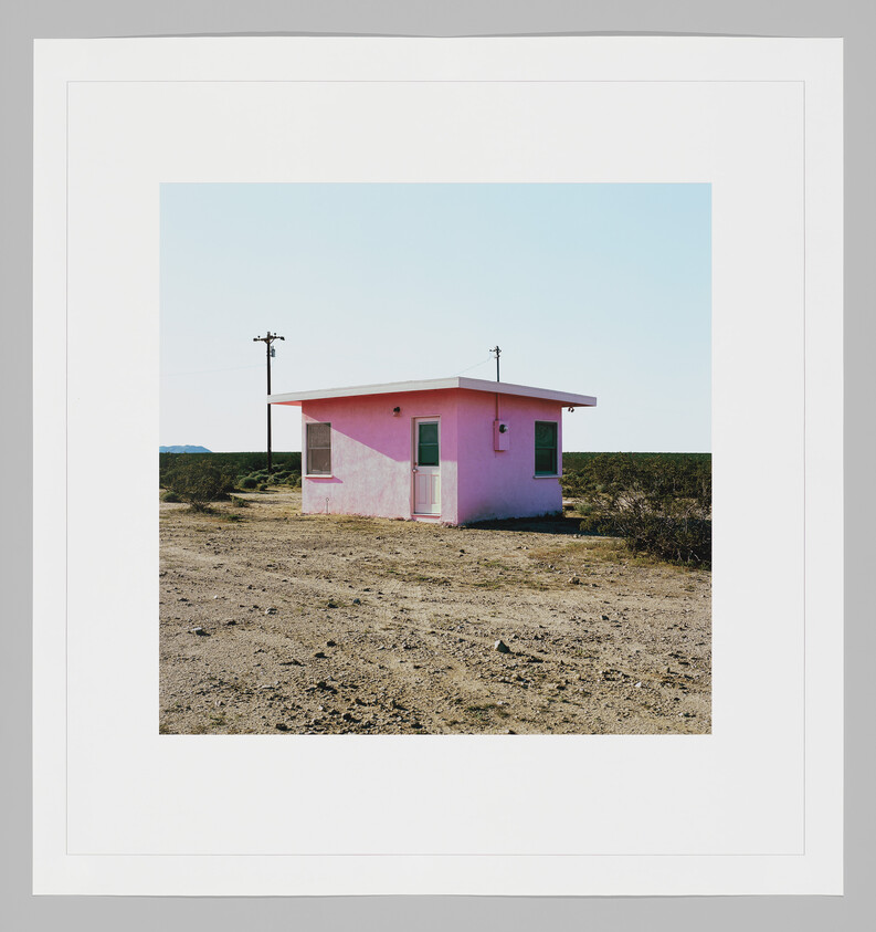 A small pink house stands alone in a barren desert landscape near two utility poles.
