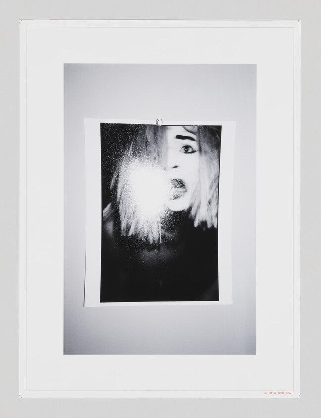 A black and white photograph within a white mat and frame, depicting a person's face partially obscured by a bright light flare. The person appears to be shouting or singing, and the image captures a dynamic, high-contrast moment. The photograph is mounted on a wall with a visible shadow around the frame, indicating it is being displayed in a gallery or similar setting.