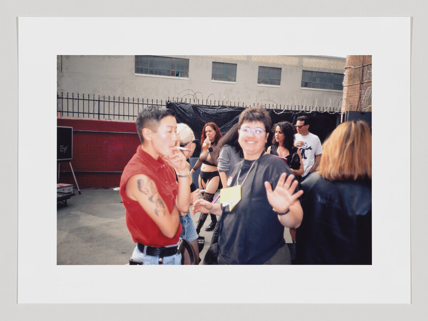 A candid photo of people at an outdoor event, with a person in a red sleeveless top smoking and another person in a black shirt smiling and waving at the camera. Other attendees are visible in the background.
