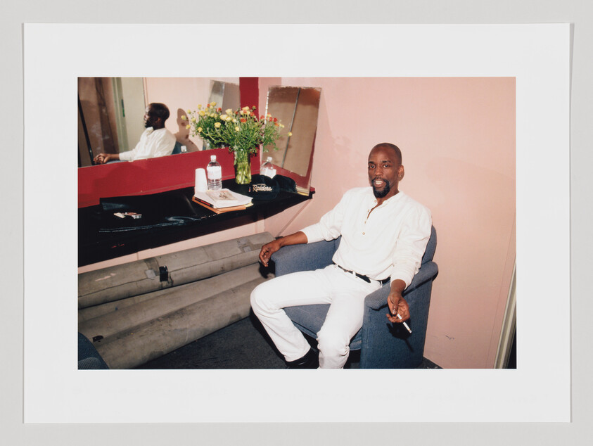 Man in white outfit sitting in a dressing room, holding a cigarette, with flowers on the table.