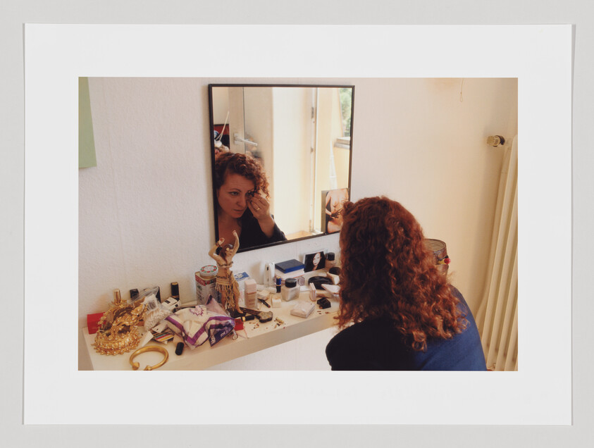 A curly, red-headed woman sits at a messy vanity doing her make-up