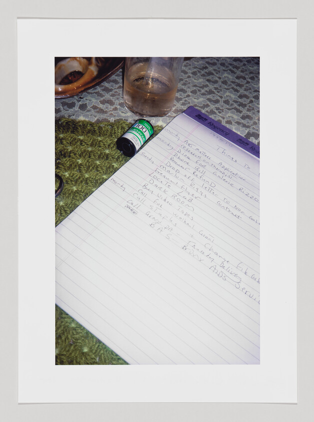 A notepad with a to-do list sitting on a lace-covered table with a roll of film and class next to it.